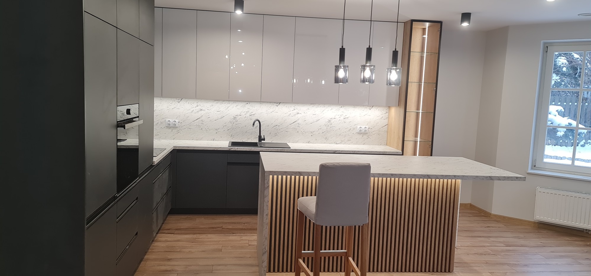 Nowoczesna kuchnia z wyspą, fronty szafek w kolorze białym i grafitowym, marmurowy blat, oświetlenie LED pod szafkami i wyspą, trzy lampy wiszące nad wyspą, widok na zaśnieżony ogród przez okno.