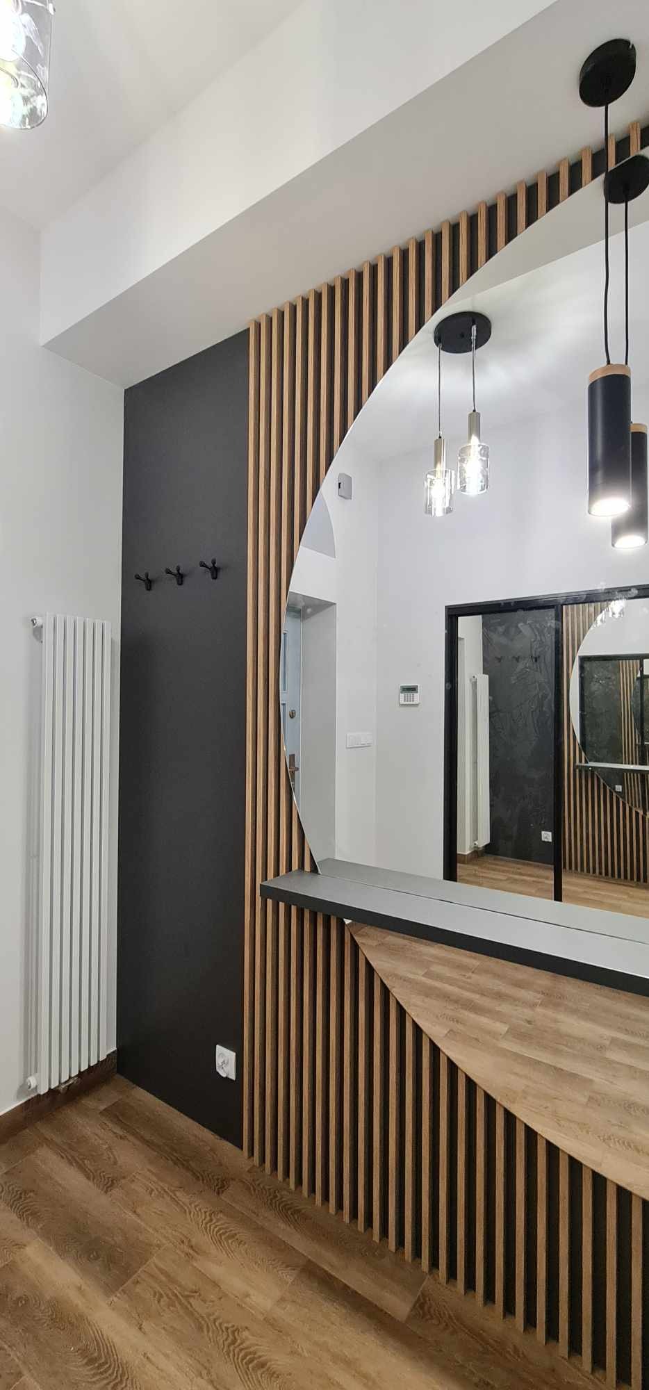 Nowoczesny przedpokój z drewnianymi lamelami na ścianie, owalnym lustrem i czarną ścianą z wieszakami, oświetlenie wiszące.