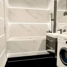 Nowoczesna łazienka z podświetlanymi białymi płytkami imitującymi marmur, czarną armaturą, umywalką z szufladami w kolorze grafitowym i pralką.