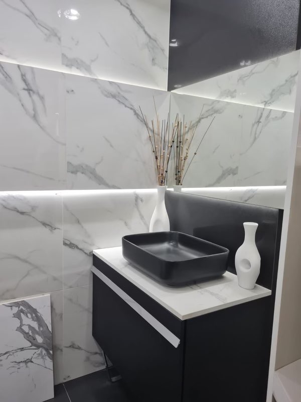 Nowoczesna łazienka z czarną, kwadratową umywalką nablatową i białymi, marmurkowymi płytkami podświetlonymi LED, uzupełniona dekoracyjnymi wazonami i suchymi gałązkami.