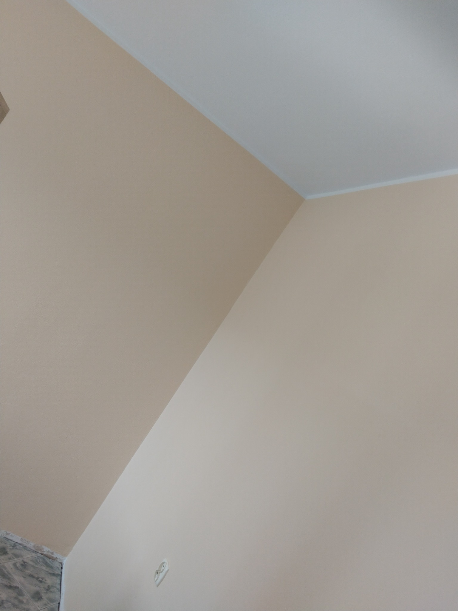 Ściana pomalowana na jasny beż w rogu pokoju, widoczny fragment sufitu z listwą wykończeniową i gniazdko elektryczne.