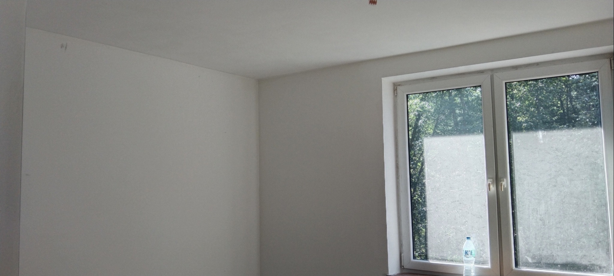 Wnętrze pokoju po malowaniu na biało, widoczne ślady po taśmie malarskiej na suficie i niedokładności przy oknie, butelka wody na parapecie.