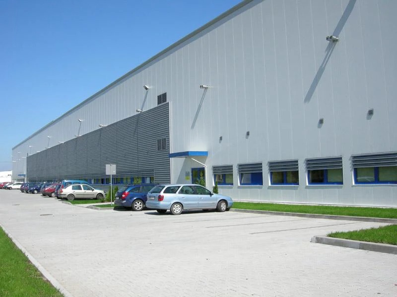 Zewnętrzny widok dużego, nowoczesnego magazynu z białymi ścianami panelowymi i szarym pasem, zaparkowane samochody wzdłuż budynku, błękitne ramy okienne.