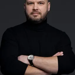 Portret mężczyzny w czarnym golfie z założonymi rękami i zegarkiem na nadgarstku, na ciemnoszarym tle.