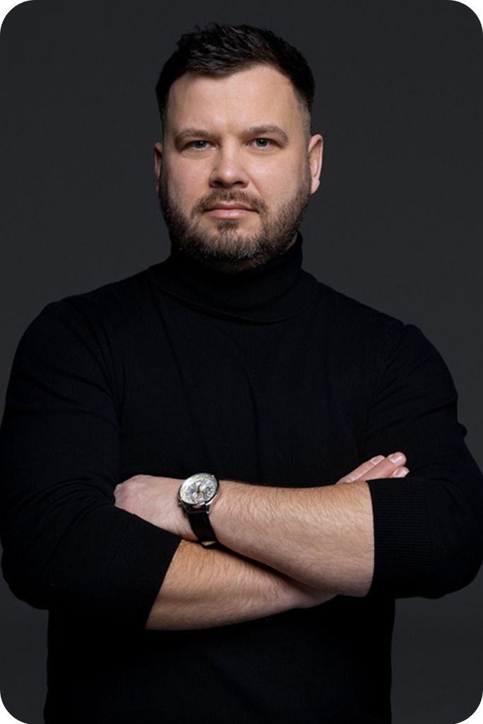 Portret mężczyzny w czarnym golfie z założonymi rękami i zegarkiem na nadgarstku, na ciemnoszarym tle.