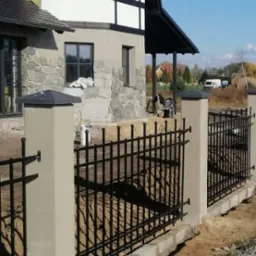 Nowoczesny dom jednorodzinny z elewacją z kamienia i białymi panelami, otoczony czarnym, metalowym ogrodzeniem na betonowej podmurówce, widoczne prace wykończeniowe wokół ogrodzenia.