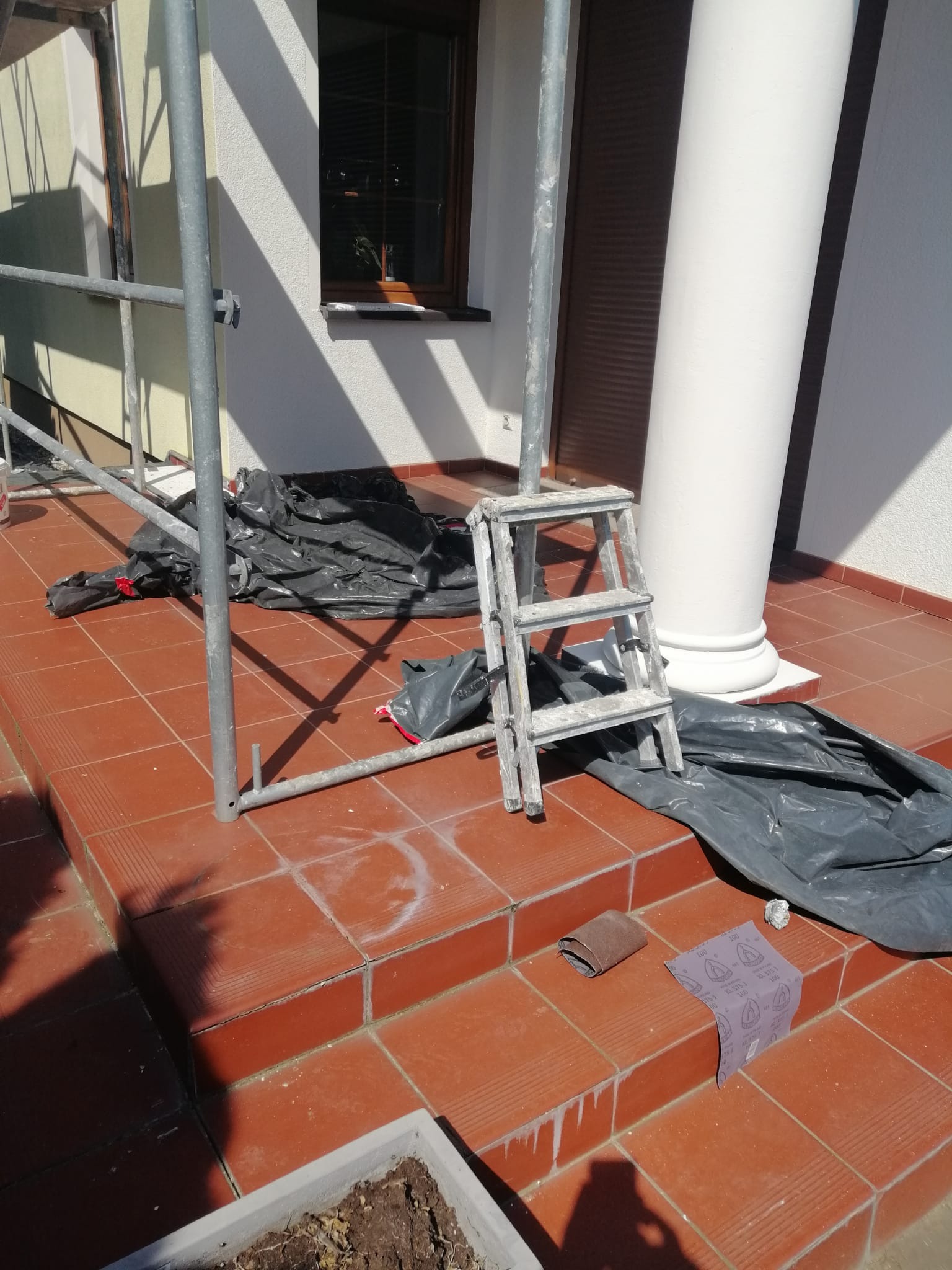 Przygotowania do prac elewacyjnych: rozstawiony rusztowanie, rozłożona folia ochronna, aluminiowa drabina oparta o kolumnę, ślady białej farby na brązowej terakocie, papier ścierny i kostka...