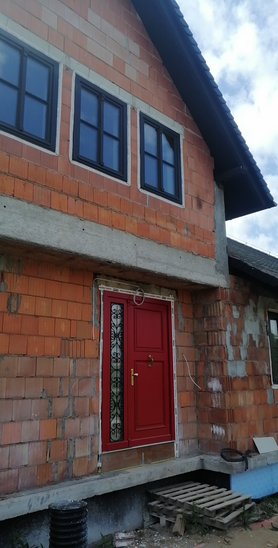 Surowa elewacja budynku w trakcie budowy, z widoczną czerwoną stolarką drzwiową i oknami z czarnymi ramami, oraz z elementami szarego betonu i pustaków ceramicznych.