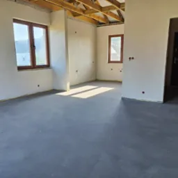 Surowe wnętrze po nałożeniu gładzi cementowej na ściany i wylewki samopoziomującej na podłodze, widoczne drewniane krokwie stropu i przygotowane otwory na gniazdka elektryczne.