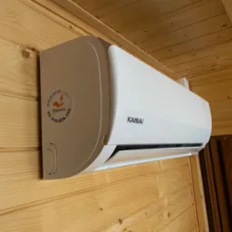 Biała klimatyzacja Kaisai zamontowana na drewnianej ścianie z widoczną naklejką serwisową Anpom z numerem telefonu.
