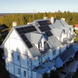 Widok z drona na budynek wielorodzinny w Bolszewie z zainstalowanymi panelami słonecznymi na dachu i nad tarasem. W tle las i inne budynki.