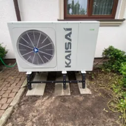 Zewnętrzna jednostka pompy ciepła Kaisai zamontowana na betonowych płytach i metalowym stelażu przed białym budynkiem z brązowym oknem i zielenią.