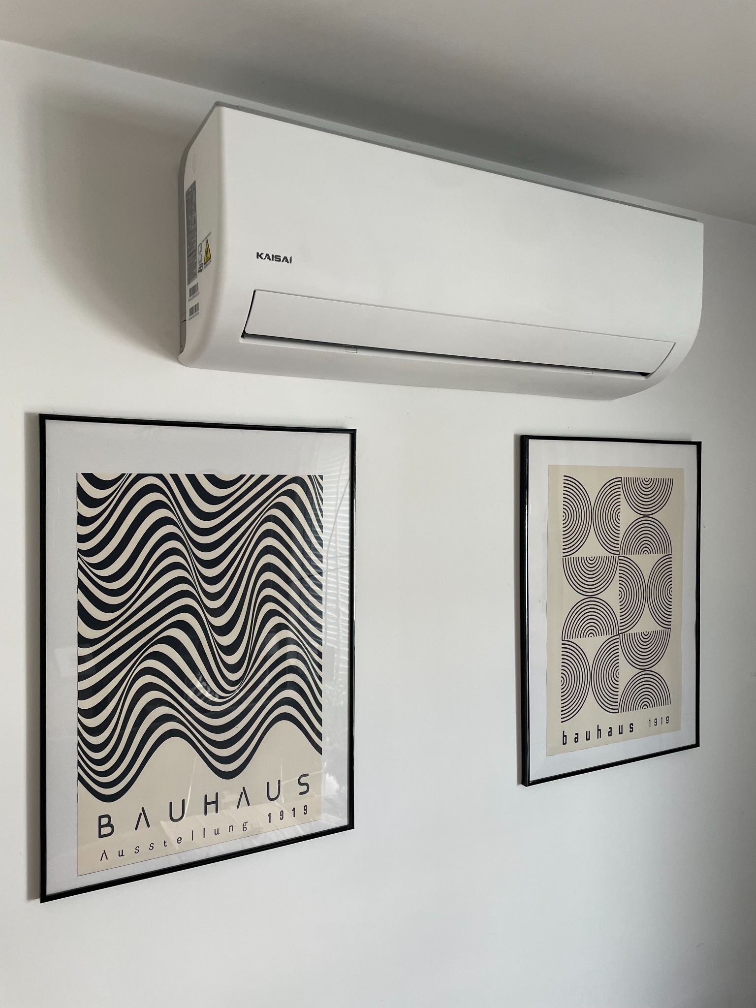 Biały klimatyzator Kaisai na ścianie, pod nim dwie oprawione grafiki Bauhaus. Minimalistyczny design wnętrza, neutralne tło.