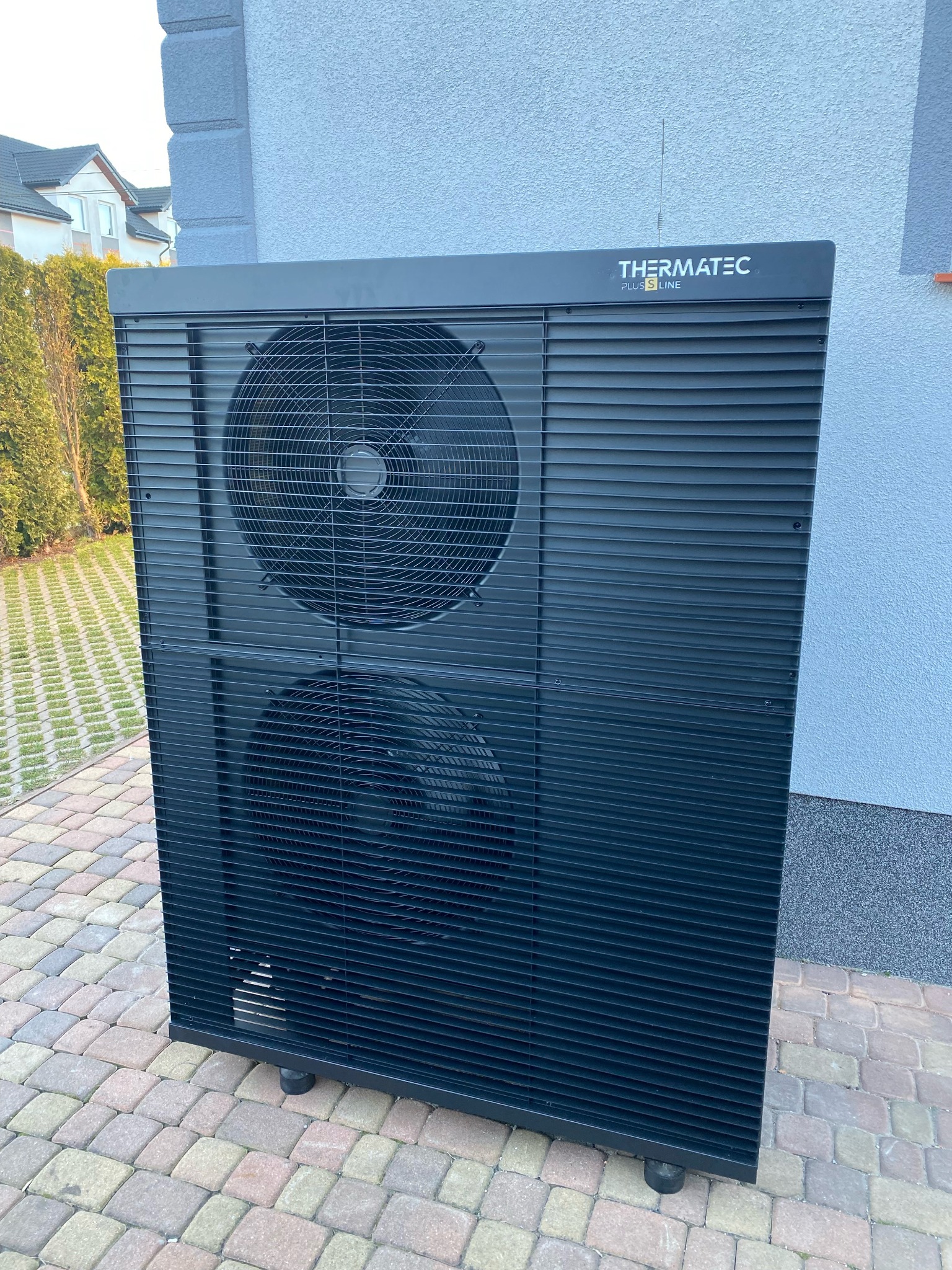 Czarna pompa ciepła Thermatec Plus S Line, zainstalowana na zewnątrz budynku, widoczna na tle kostki brukowej i elewacji. Dwa wentylatory w obudowie.