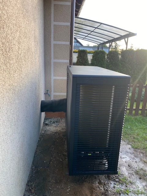 Zamontowana pompa ciepła powietrze-woda przy ścianie budynku z widocznym przyłączem. Tło: ogród i zadaszenie. Ujęcie z boku, w pochmurny dzień.