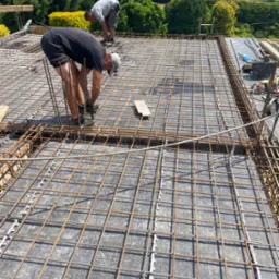 Dwóch mężczyzn układa siatkę zbrojeniową na deskowaniu stropu, widoczne elementy dystansowe z tworzywa, w tle zieleń drzew.