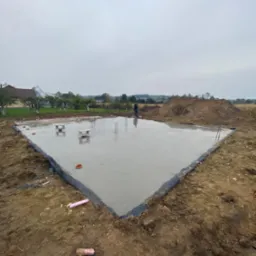 Świeżo wylany, trójkątny fundament z wystającymi prętami zbrojeniowymi, zabezpieczony folią kubełkową, widok z góry na plac budowy w pochmurny dzień.