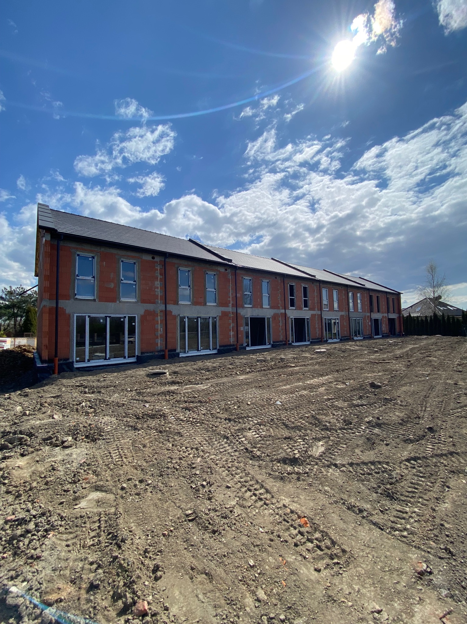 Szeregówka w stanie surowym otwartym, z widocznymi oknami i dachem, na tle błękitnego nieba z chmurami. Teren wokół budynku przygotowany pod dalsze prace.