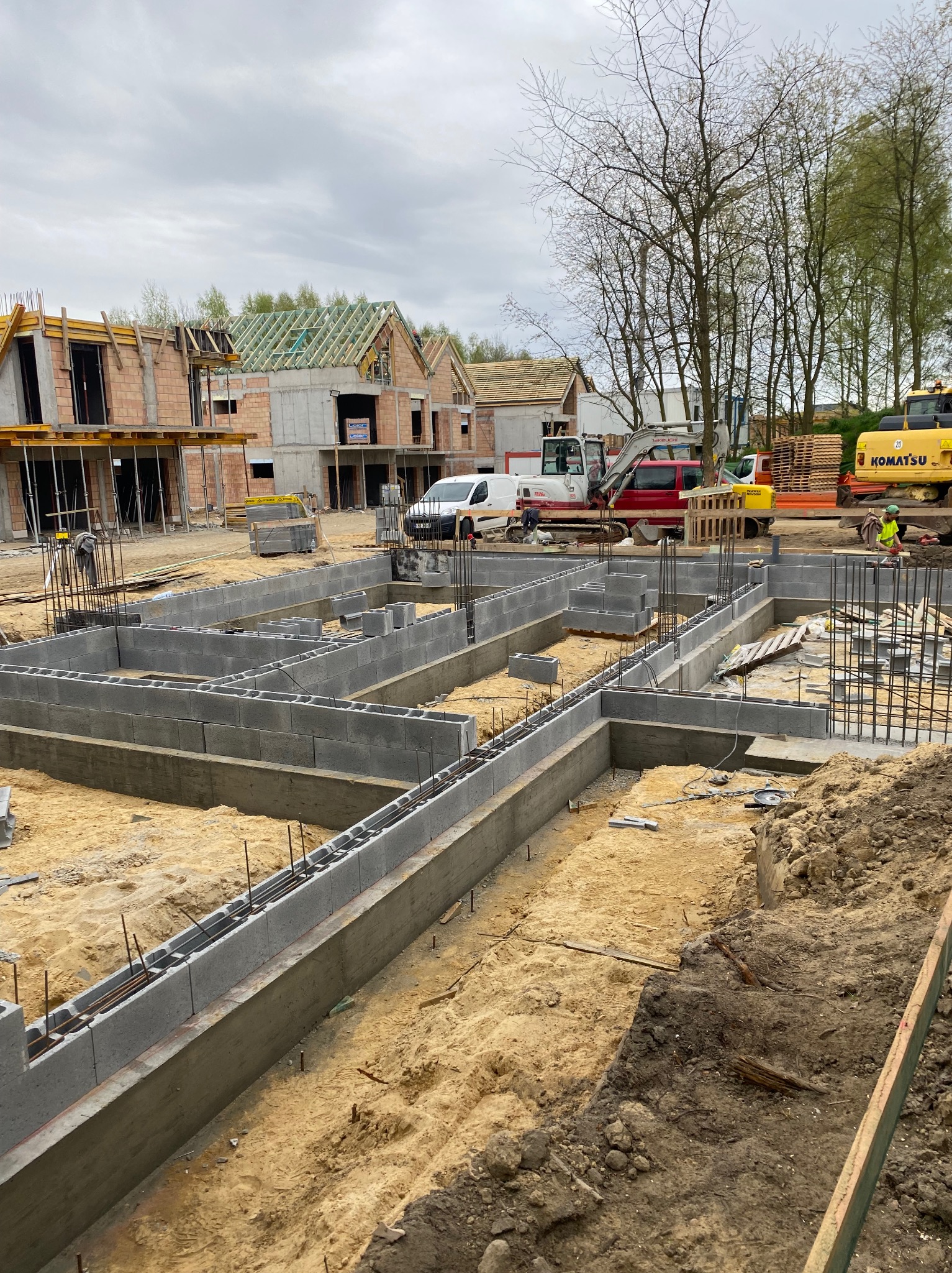 Fundamenty zbrojone pod budowę domów jednorodzinnych w Krakowie. Widoczne bloczki betonowe, stalowe pręty zbrojeniowe i wykopane wykopy. W tle budynki w trakcie budowy.