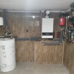 Kotłownia z białym zasobnikiem ciepłej wody owiniętym folią, wiszącym kotłem gazowym Buderus, czerwonym zbiornikiem wyrównawczym i szarymi rurami izolowanymi, na tle kafelkowanych ścian.