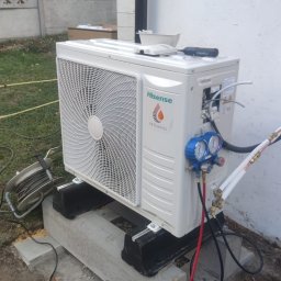 SYNERGA INSTALACJE - Zewnętrzna jednostka klimatyzacji Hisense zamontowana na betonowych blokach, podłączona do pompy próżniowej i manometrów, na tle białej ściany i betonowego ogrodzenia.