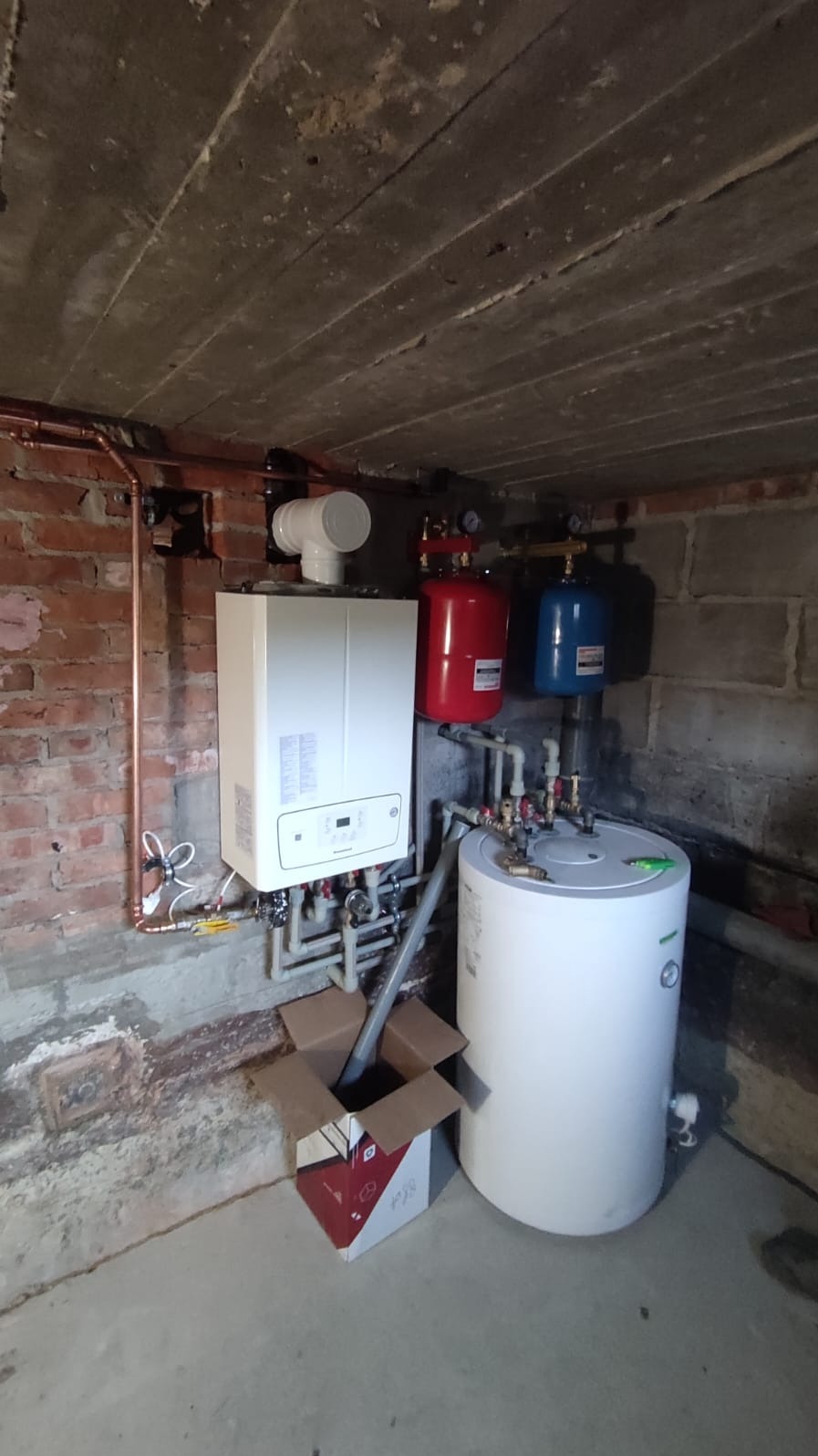 Nowy, biały piec grzewczy kondensacyjny zamontowany na ceglanej ścianie w piwnicy, z widocznymi rurami miedzianymi i plastikowymi, obok stoi biały zasobnik na wodę i otwarty karton.
