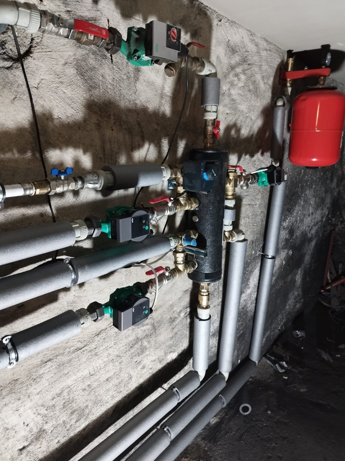 Rozbudowana instalacja grzewcza z licznymi rurami izolowanymi termicznie, zaworami, pompami obiegowymi i zbiornikiem wyrównawczym na tle szarej, surowej ściany.