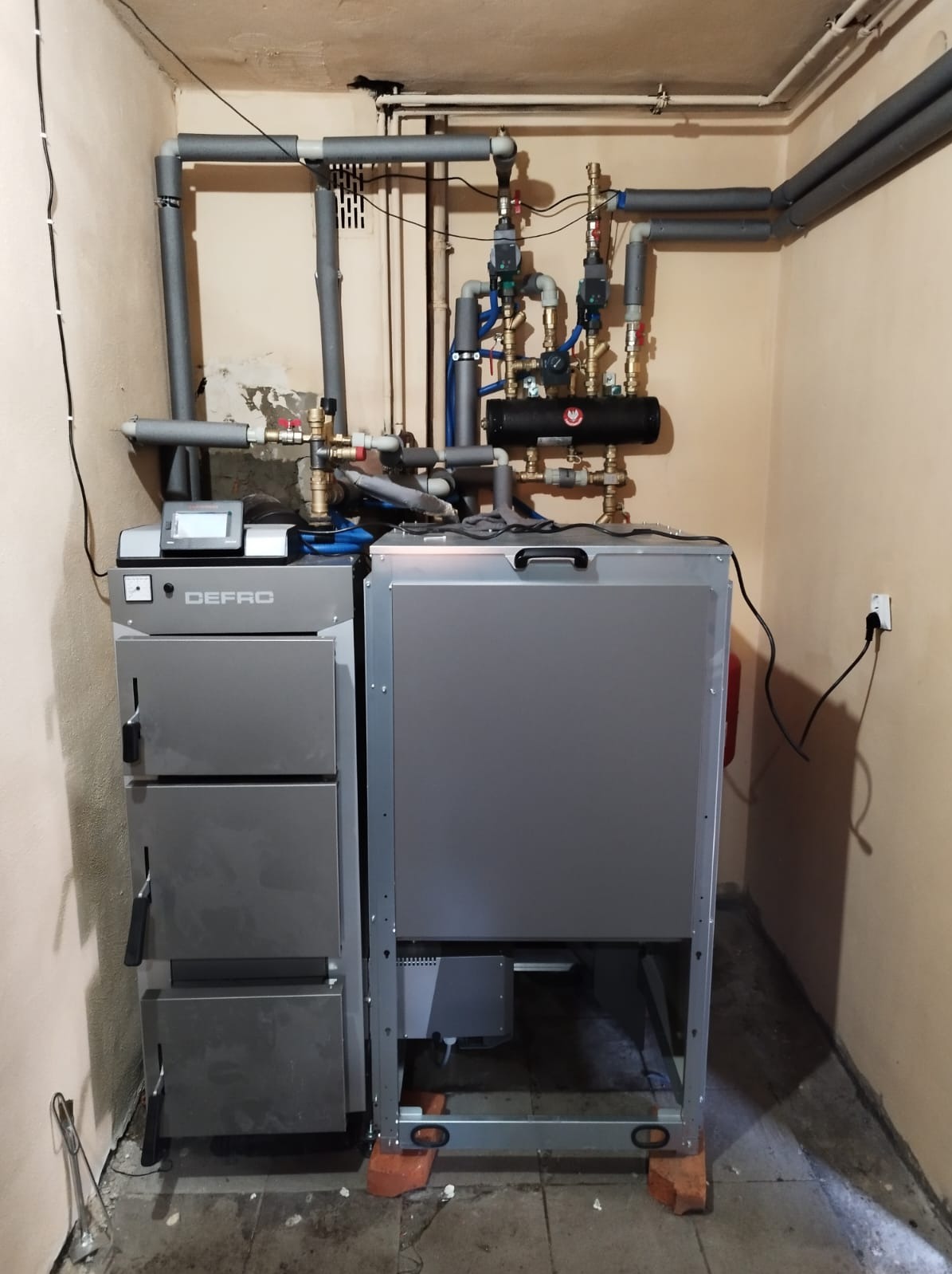 Szary piec CO marki Defro z podajnikiem, widoczne rury instalacji grzewczej z izolacją termiczną oraz armatura, zamontowany w kotłowni o beżowych ścianach.