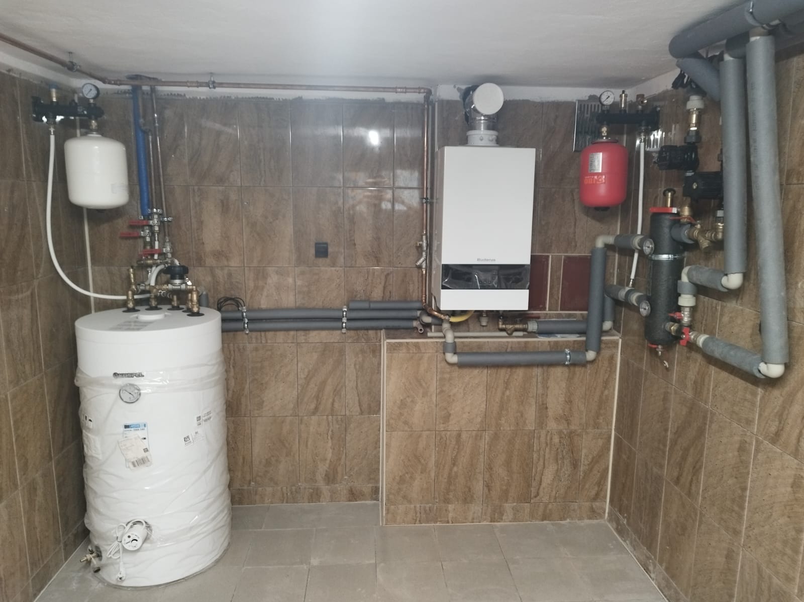 Kotłownia z białym zasobnikiem ciepłej wody owiniętym folią, wiszącym kotłem gazowym Buderus, czerwonym zbiornikiem wyrównawczym i szarymi rurami izolowanymi, na tle kafelkowanych ścian.