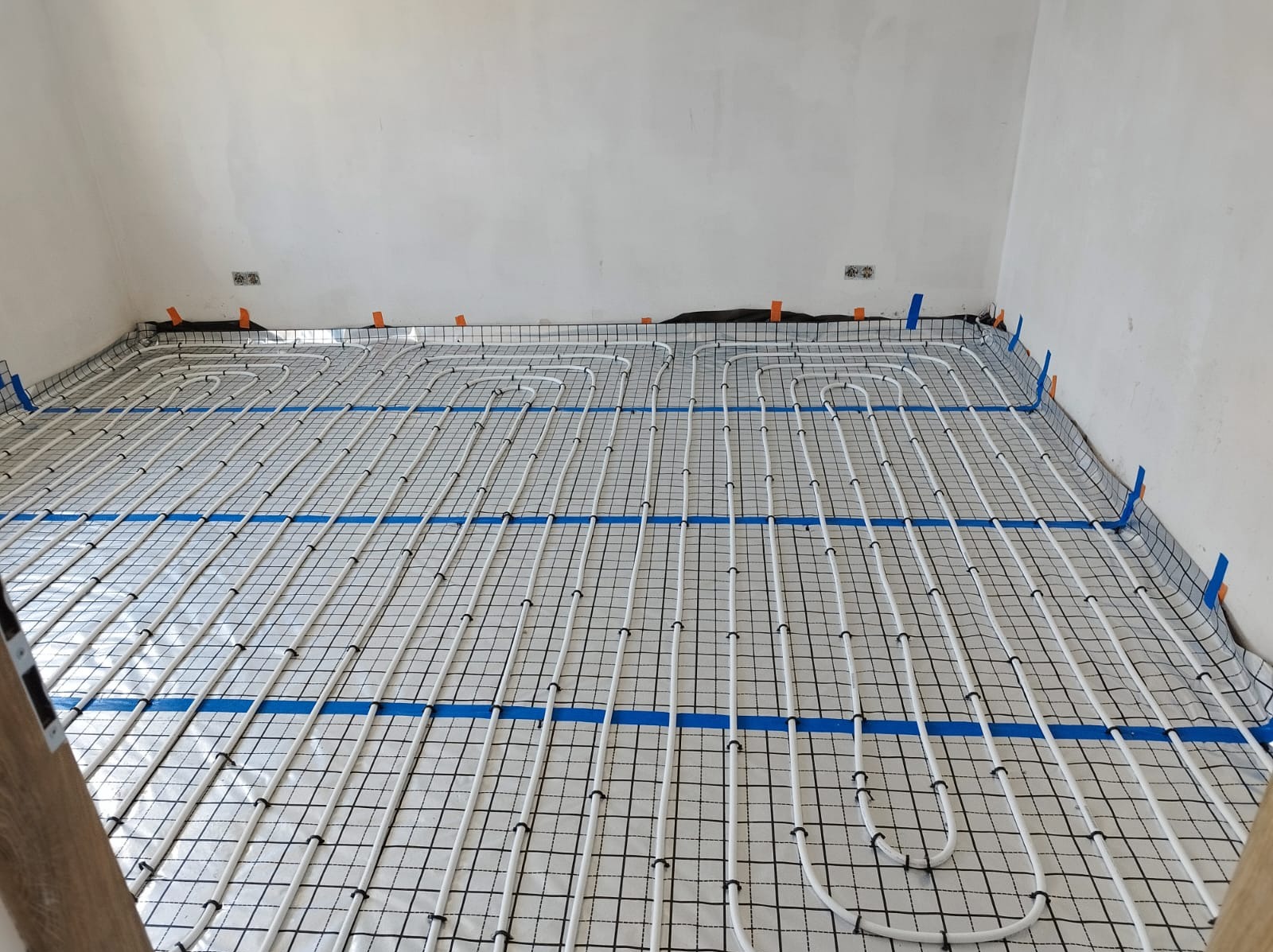 Białe rury ogrzewania podłogowego ułożone na czarnej siatce z folią aluminiową, przymocowane niebieską taśmą do ścian w surowym wnętrzu.