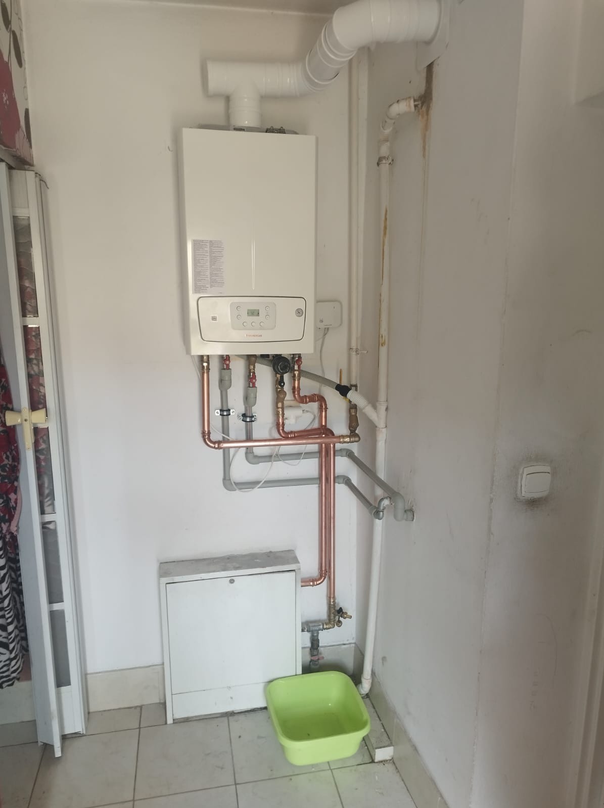 Biały piec gazowy dwufunkcyjny z podłączeniami miedzianymi i plastikowymi rurami, zamontowany na ścianie nad skrzynką i zielonym pojemnikiem.