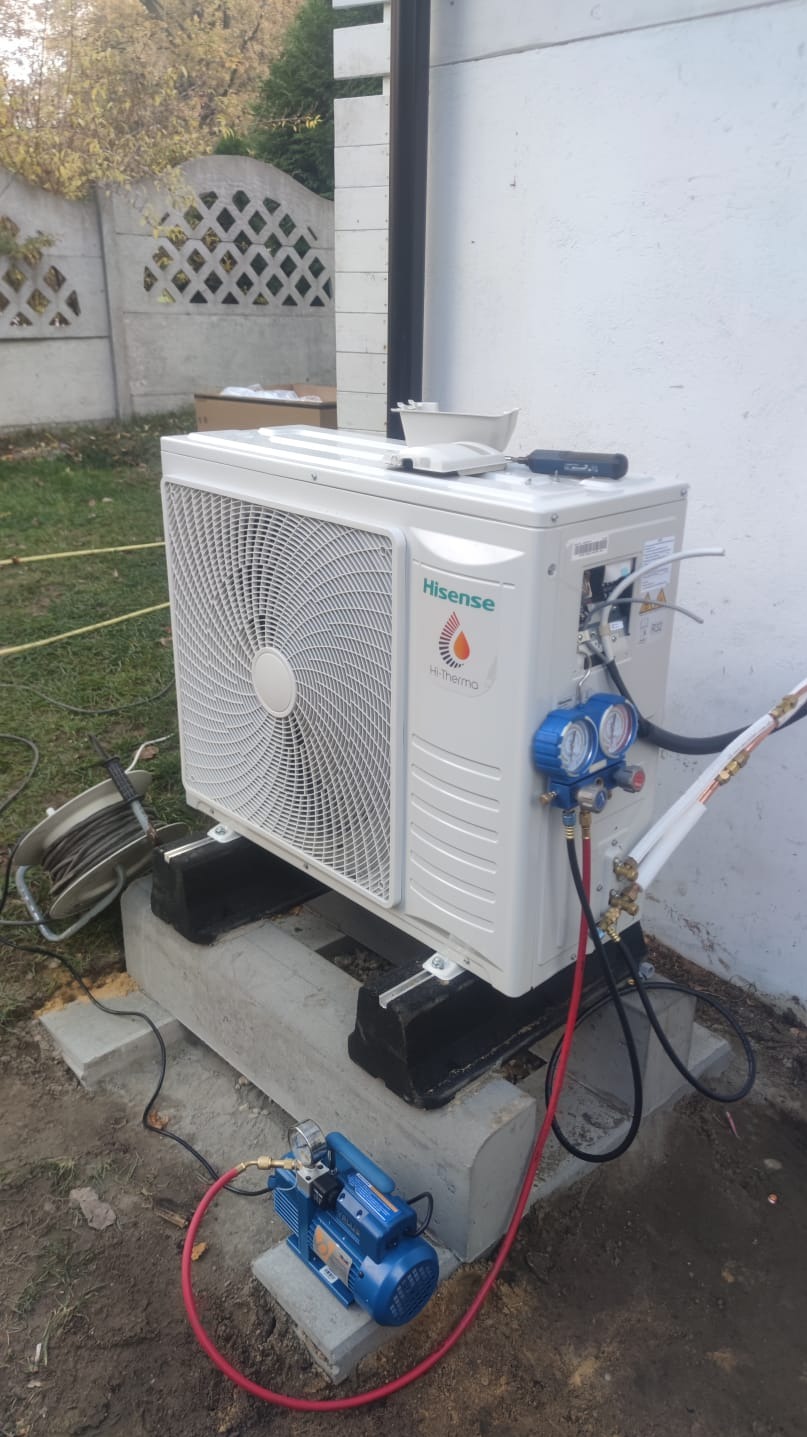 Zewnętrzna jednostka klimatyzacji Hisense zamontowana na betonowych blokach, podłączona do pompy próżniowej i manometrów, na tle białej ściany i betonowego ogrodzenia.