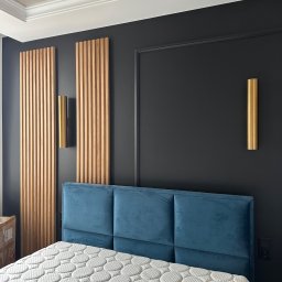 FPHU „MARKUZ” Marcin Kuzara - Nowoczesna sypialnia z łóżkiem z niebieskim, aksamitnym zagłówkiem, drewnianymi panelami ściennymi i złotymi lampami na czarnej ścianie z dekoracyjną sztukaterią.