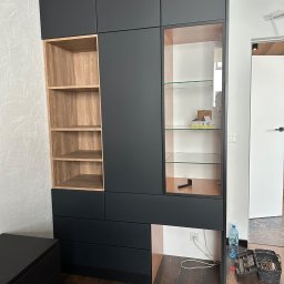 FPHU „MARKUZ” Marcin Kuzara - Czarna szafa wnękowa z drewnianymi półkami i szklaną witryną, widoczne narzędzia i butelka wody na podłodze.