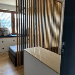 Drewniana ścianka działowa z pionowych listew w kolorach naturalnego drewna i czerni, oddzielająca przestrzeń sypialną od biurowej, widoczna zza uchylonych drzwi.