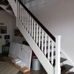 FPHU „MARKUZ” Marcin Kuzara - Drewniane schody z białymi balustradami i ciemnobrązowymi stopniami, widoczne materiały budowlane i metalowe wiadro na podłodze z płytek.