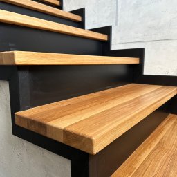 FPHU „MARKUZ” Marcin Kuzara - Nowoczesne schody z drewnianymi stopniami o ciepłym odcieniu i czarną metalową konstrukcją, widoczne z dołu, na tle szarej, surowej ściany.