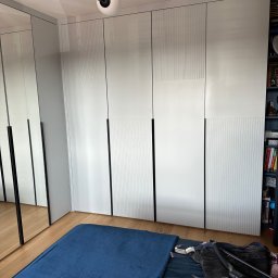 FPHU „MARKUZ” Marcin Kuzara - Biała szafa wnękowa z subtelnym wzorem pionowych linii na frontach, czarne, minimalistyczne uchwyty, odbicie w lustrzanych drzwiach szafy, widoczne elementy wyposażenia sypialni.