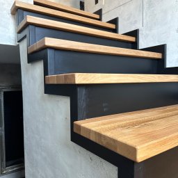FPHU „MARKUZ” Marcin Kuzara - Nowoczesne schody z drewnianymi stopniami i czarną metalową konstrukcją, betonowa ściana w tle, widok z dołu.