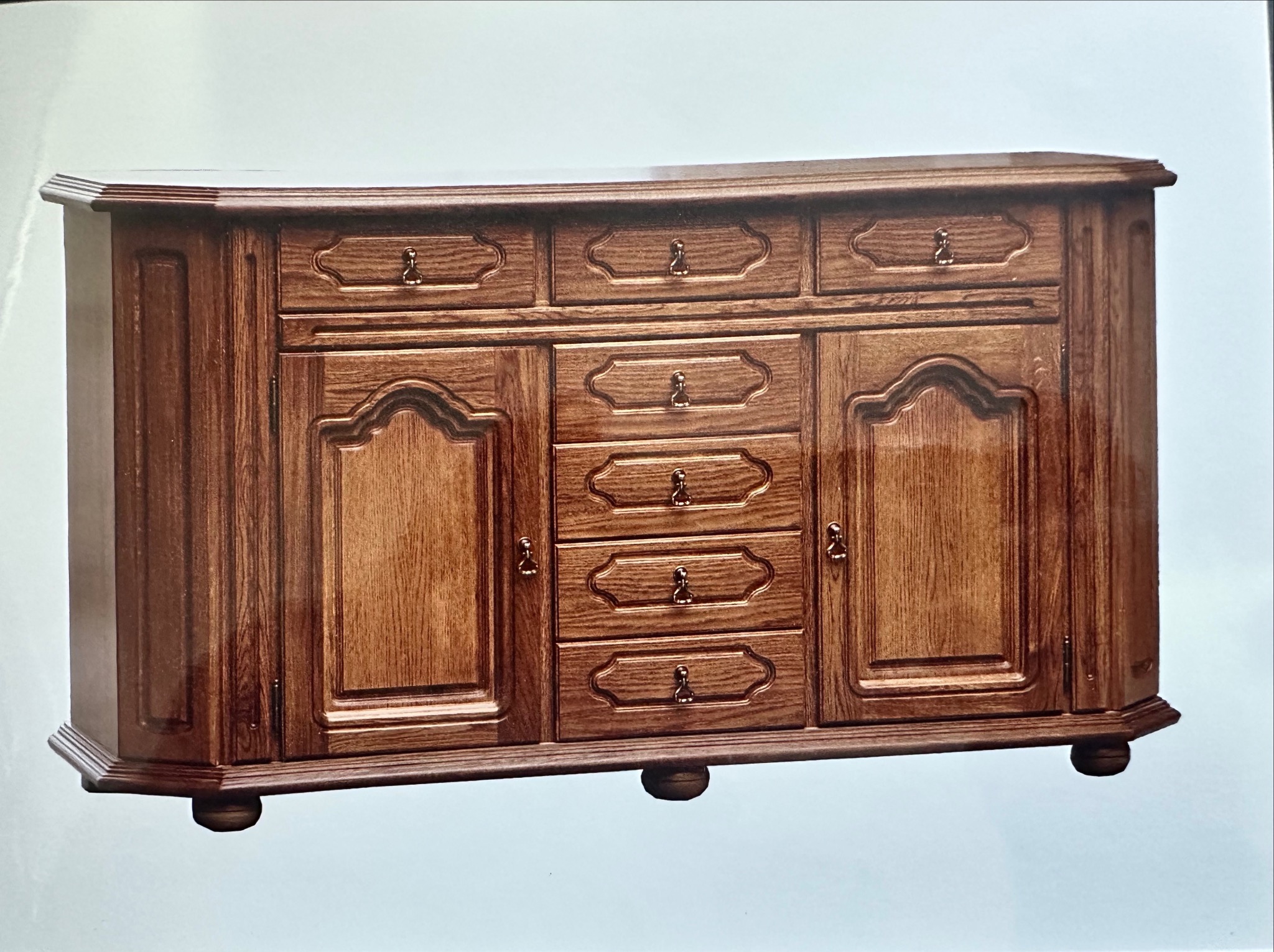 Klasyczna, fornirowana komoda z litego drewna dębowego, z rzeźbionymi frontami szuflad i drzwiczek, na niskich, okrągłych nóżkach. Ciepłe oświetlenie podkreśla rysunek słojów.