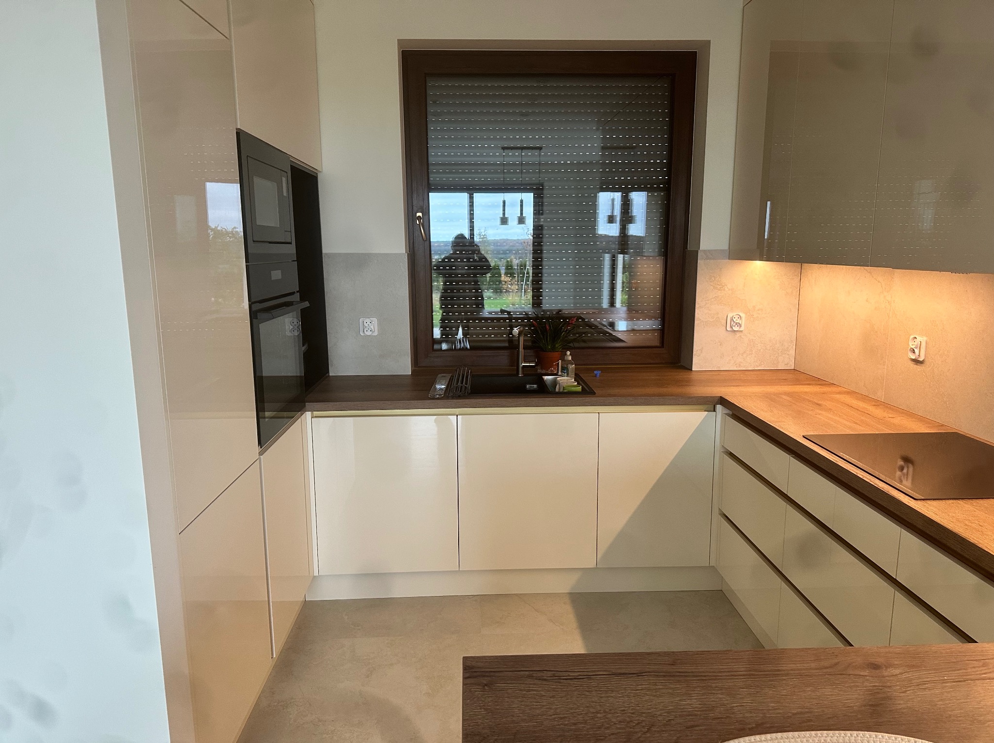 Nowoczesna kuchnia z białymi, lakierowanymi frontami, drewnianym blatem i oknem z roletami, w którym odbija się sylwetka osoby na tle zieleni.