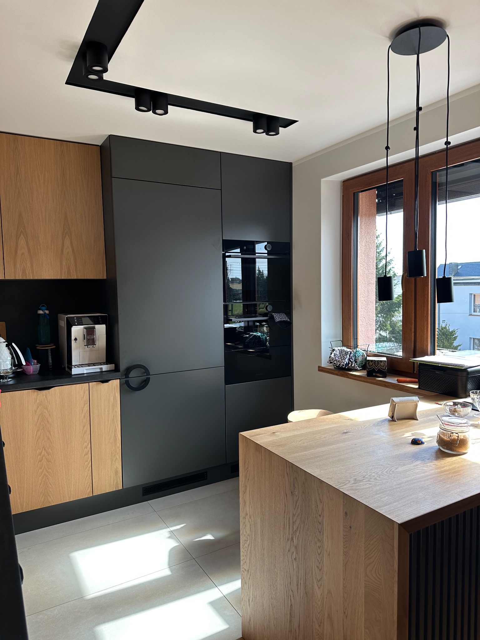 Nowoczesna kuchnia z drewnianymi i grafitowymi frontami, wyspą z blatem z jasnego drewna oraz oknem z widokiem na zieleń.