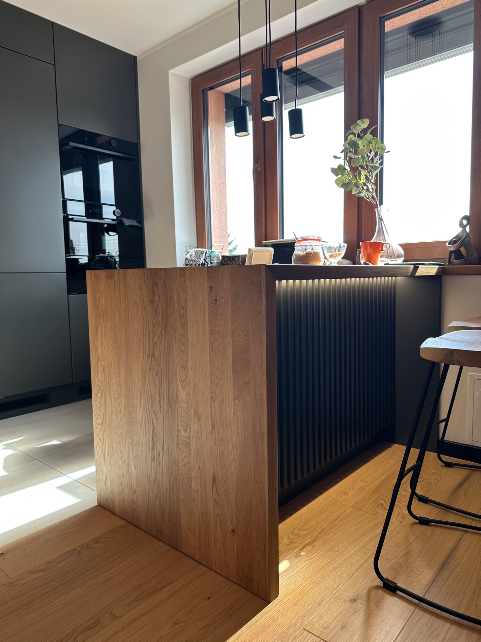 Wyspa kuchenna z drewnianym frontem i podświetleniem LED, widok na nowoczesną kuchnię z czarnymi szafkami i drewnianymi oknami, na blacie wazon z zielonymi gałązkami i inne dekoracje.