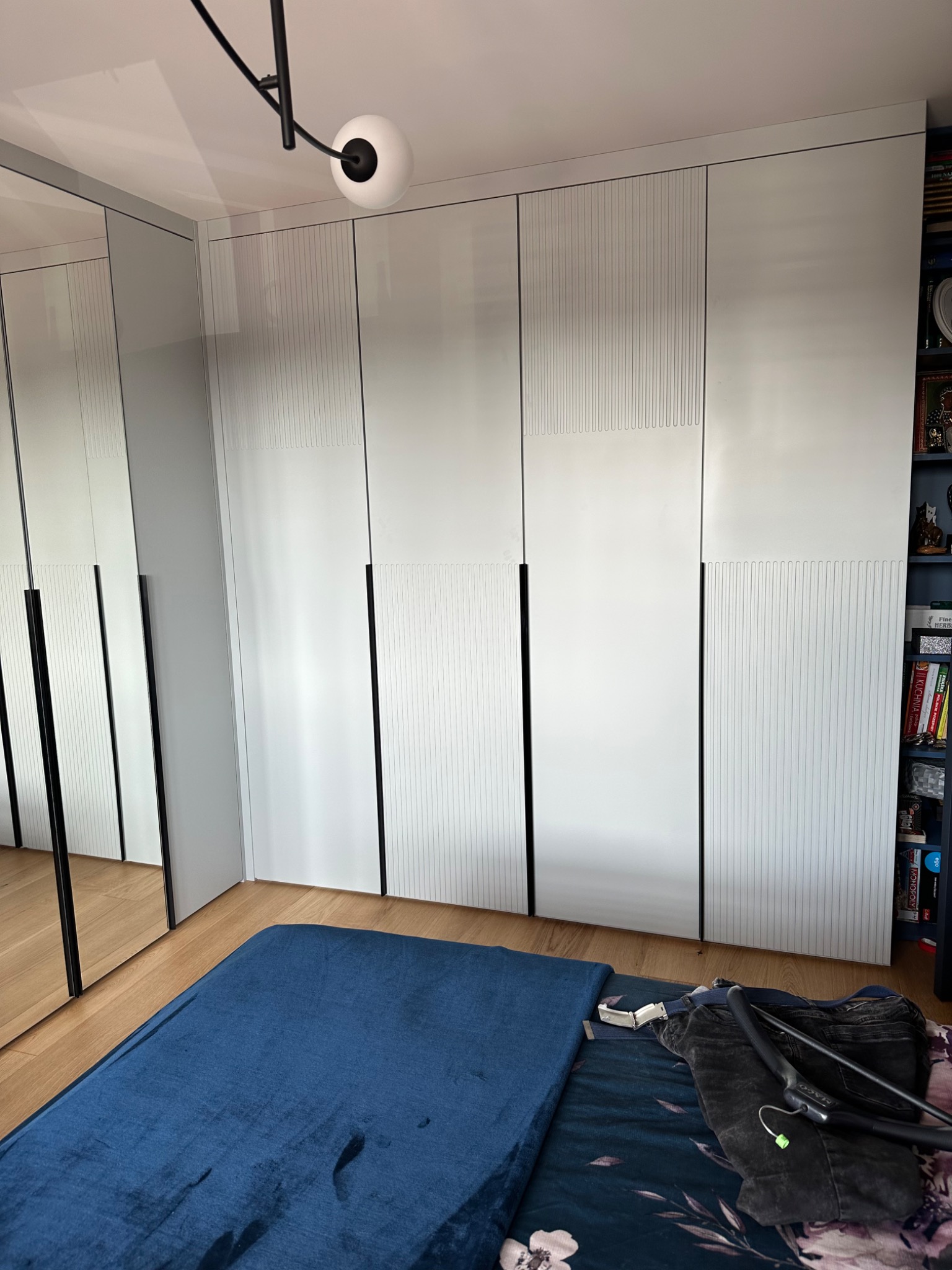 Biała szafa wnękowa z subtelnym wzorem pionowych linii na frontach, czarne, minimalistyczne uchwyty, odbicie w lustrzanych drzwiach szafy, widoczne elementy wyposażenia sypialni.