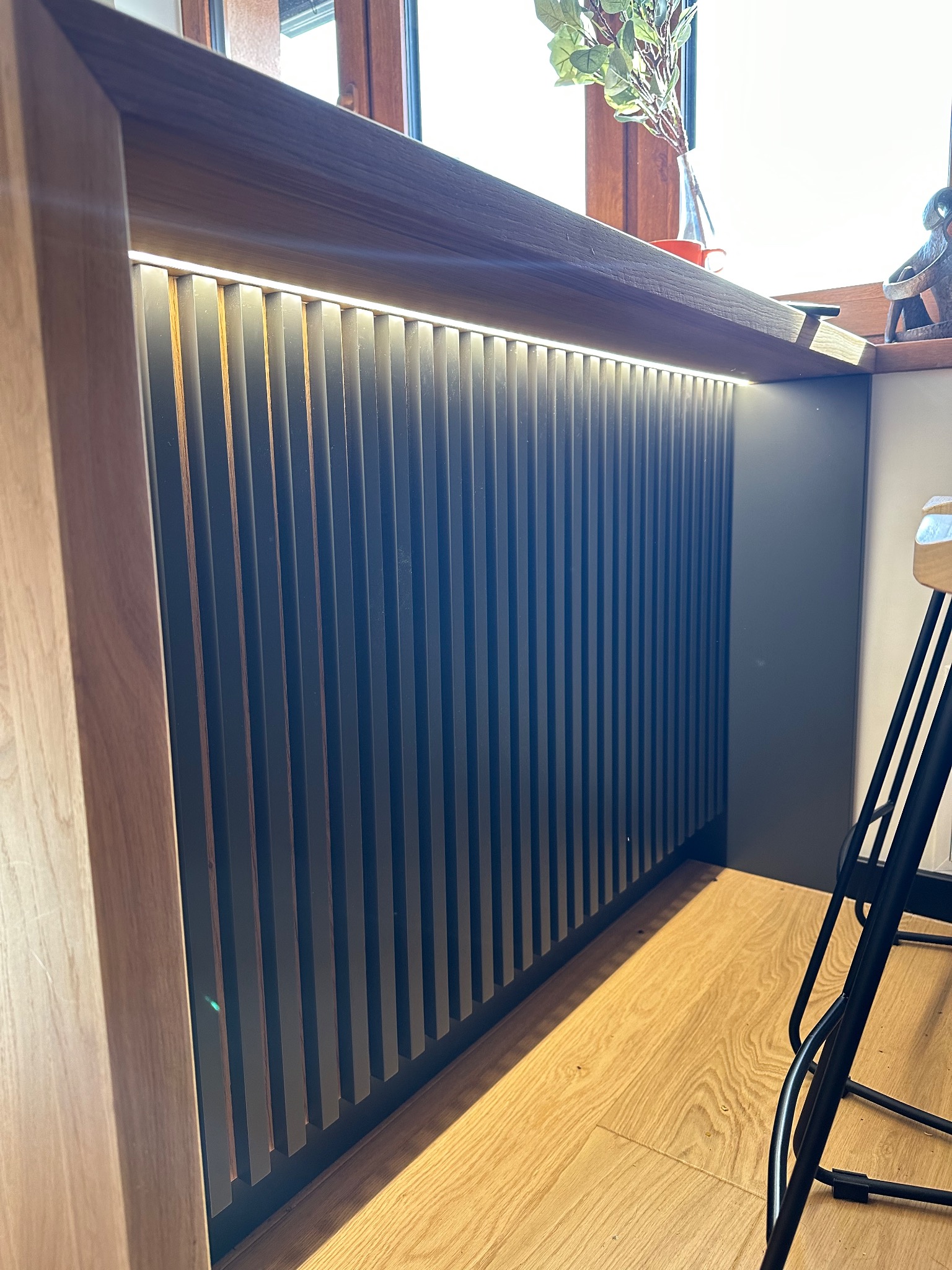 Nowoczesna zabudowa kuchenna z pionowymi lamelami podświetlonymi listwą LED, drewniany blat i fragment krzesła barowego na parkiecie.