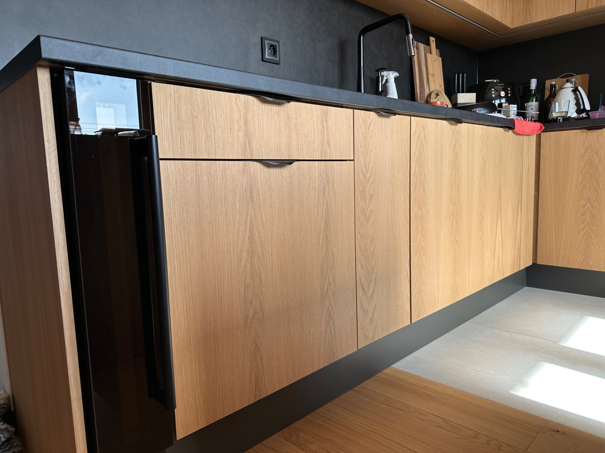 Fragment zabudowy kuchennej z frontami z jasnego drewna z widocznymi słojami, czarny blat i cokół, nowoczesne uchwyty, widoczny sprzęt AGD i akcesoria kuchenne.