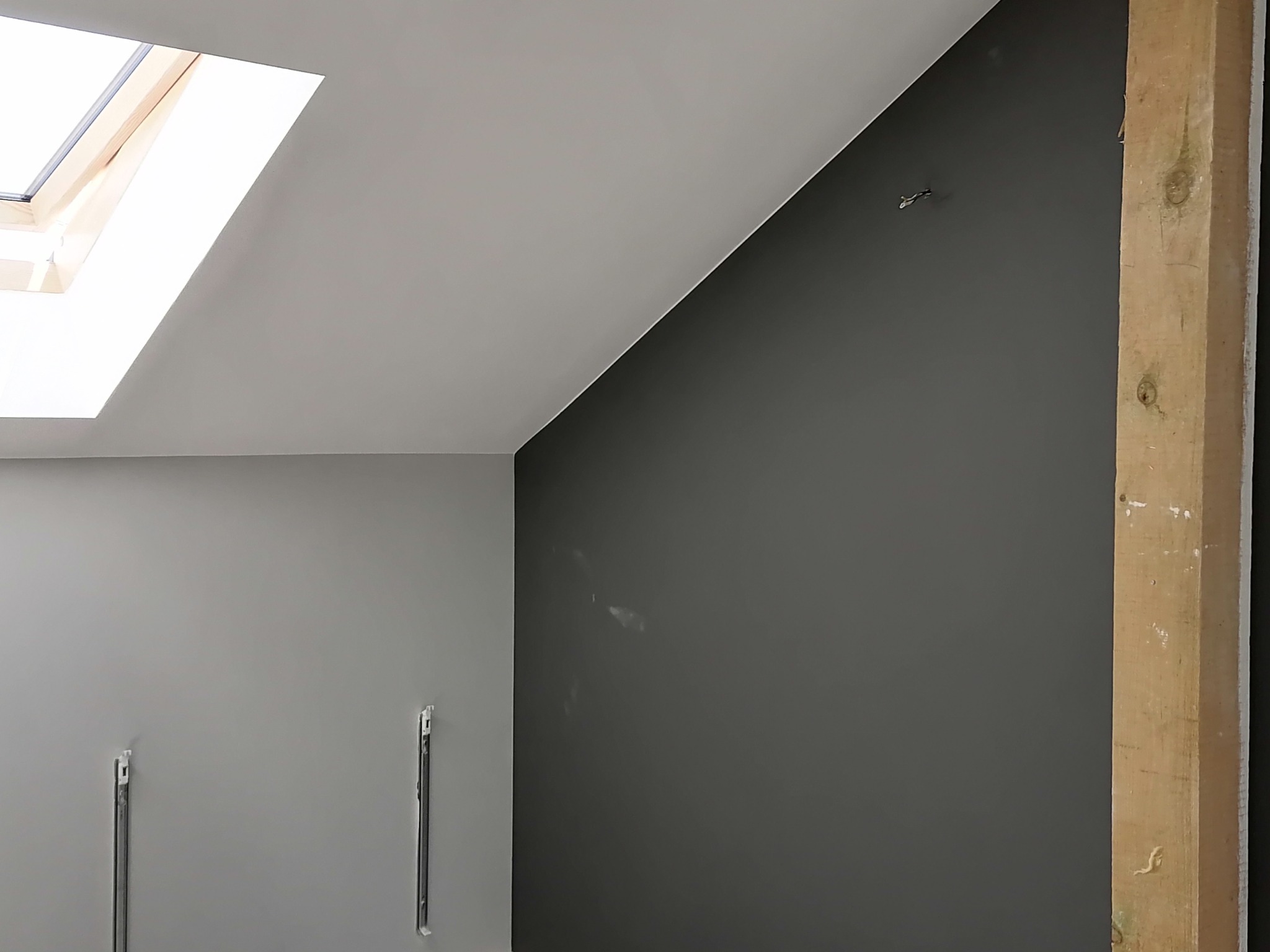 Świeżo pomalowane wnętrze poddasza w odcieniach szarości, z drewnianym słupem konstrukcyjnym i oknem dachowym. Na ścianie widoczne ślady szpachlowania i metalowe listwy mocujące.