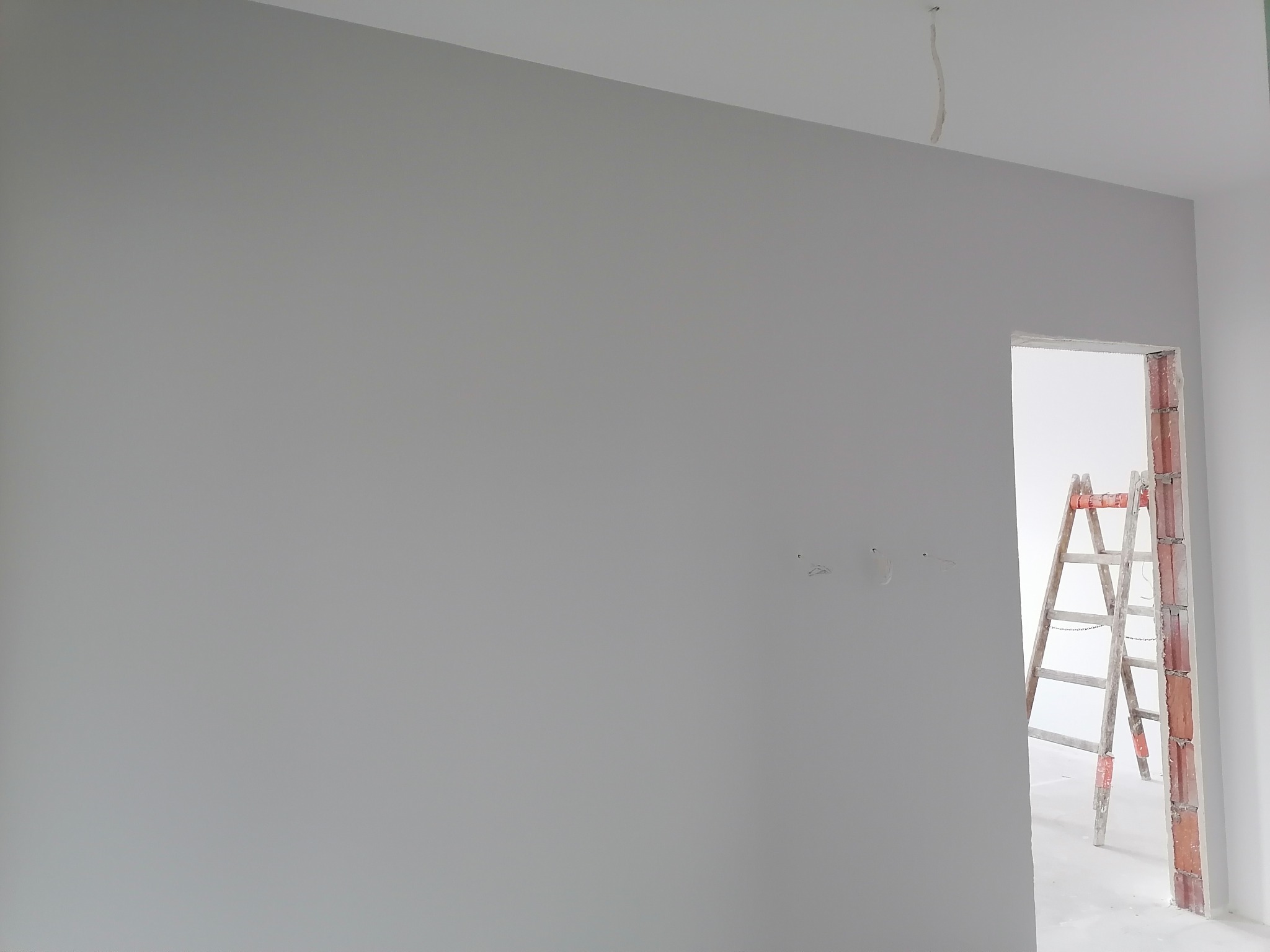 Świeżo pomalowana szara ściana z widocznymi śladami po gładzi, drabina oparta o framugę drzwi, widoczne wystające kable ze ściany.