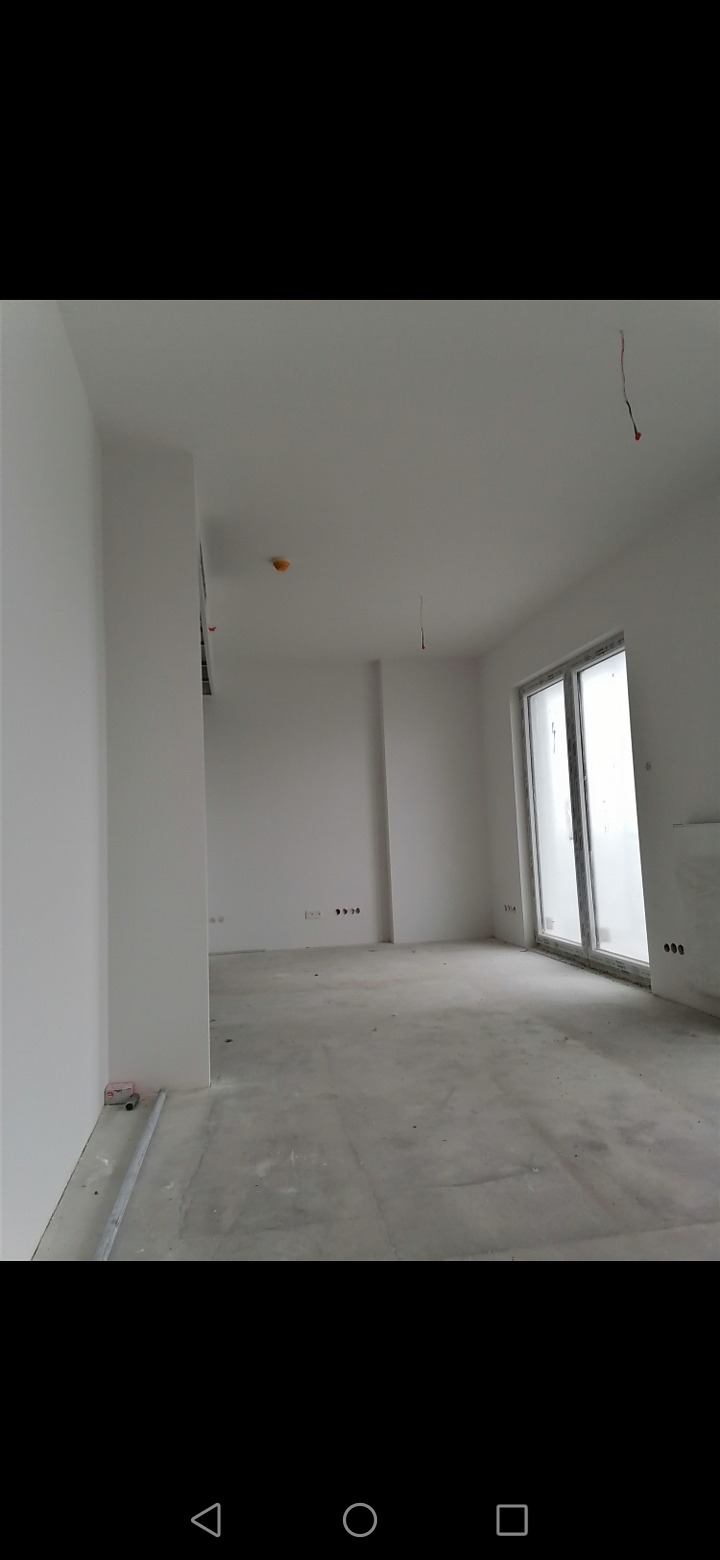 Surowe wnętrze po remoncie, widoczne świeżo pomalowane ściany na biało, betonowa podłoga, wystające kable z sufitu i niezabezpieczony czujnik dymu.