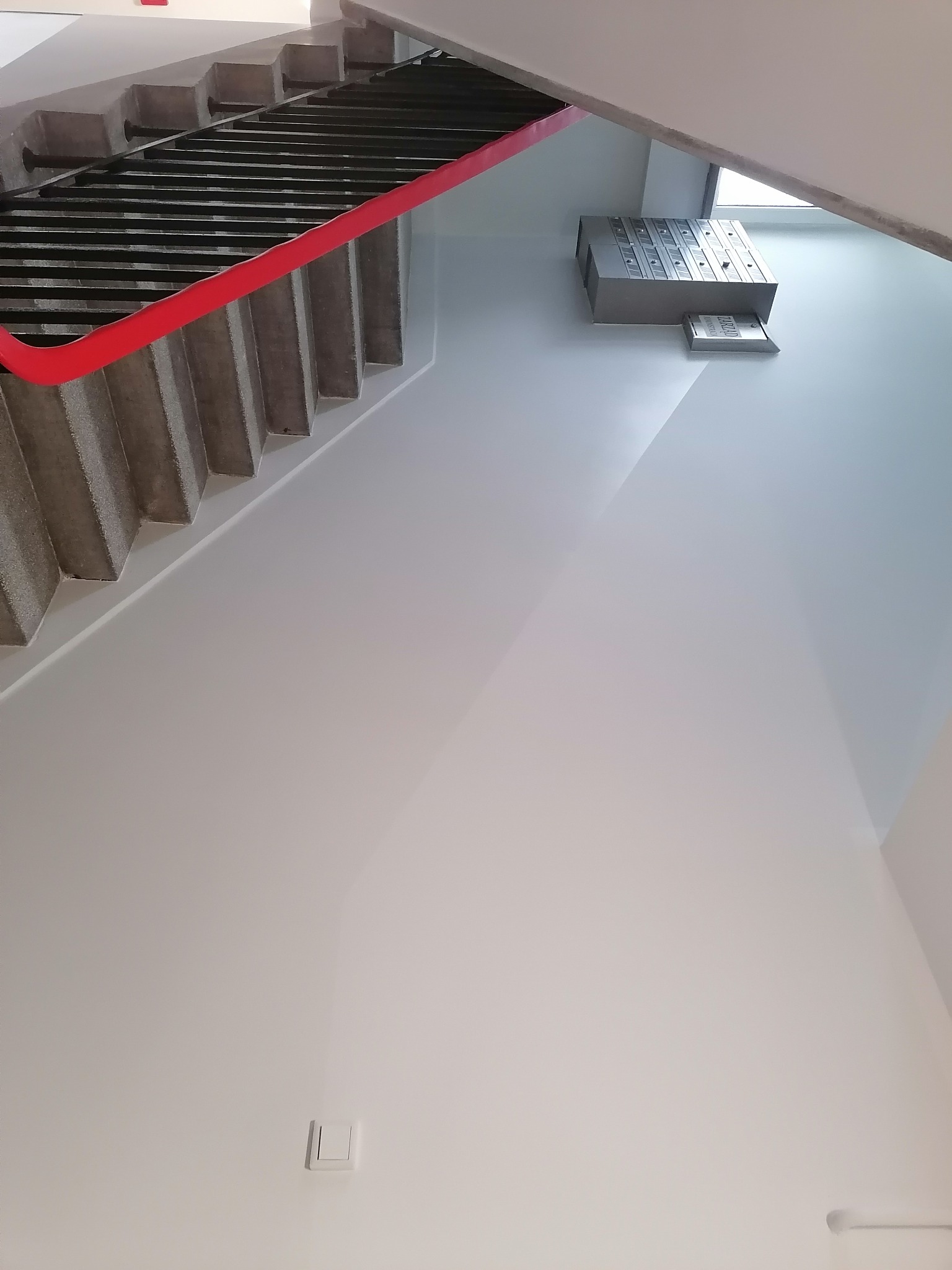 Klatka schodowa w budynku wielorodzinnym po odświeżeniu, ściany pomalowane na jasny odcień szarości, widoczne skrzynki pocztowe i włącznik światła.